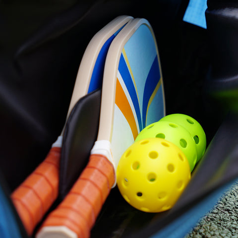 The Padel & Pickleball Bundle from Cancha includes two pickleball paddles with orange grips and two yellow pickleballs, all neatly stored inside a sleek black padel bag.