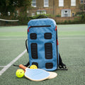 The Cancha Padel & Pickleball Bundle, including a blue backpack, a wet-dry bag, and two pickleball paddles with balls, is set on a tennis court with a brick building in the background.