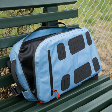 The Padel & Pickleball Bundle by Cancha, featuring a light blue bag with black accents and a red zipper pull, rests on a green bench against the backdrop of a chain-link fence. This versatile design includes modular accessories for added convenience.