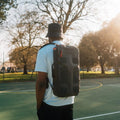 A person stands on an outdoor basketball court with trees in the background, wearing a Cancha bucket hat and backpack from the Padel & Pickleball Bundle, their modular accessories seamlessly integrated for a day of adventure.