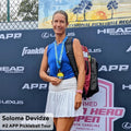 A woman in a sports outfit holds a gold medal, standing in front of a background adorned with various sports brand logos. With her Cancha Padel & Pickleball Bundle bag by her side, the text reveals her name is Solome Devidze and she is ranked #2 on the APP Pickleball Tour.