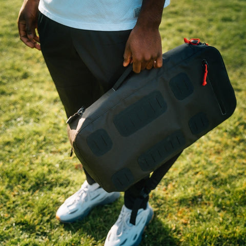 A person is standing on grass holding the Padel & Pickleball Bundle from Cancha, a black duffel bag with a red zipper pull. They are dressed in a white shirt, dark pants, and white shoes. The Cancha duffel functions as a versatile modular accessory ideal for various activities.