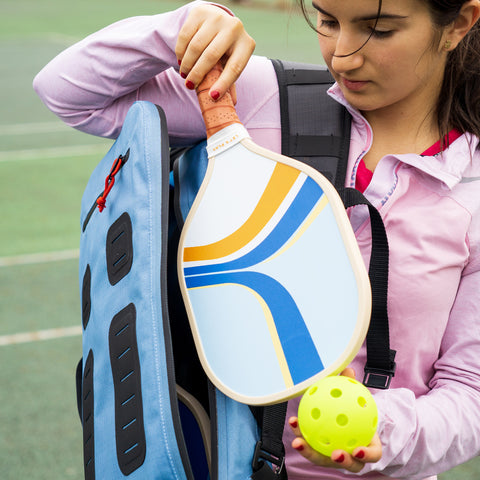 A person in a pink jacket takes the Padel & Pickleball Bundle by Cancha, including a paddle and ball, out of a blue backpack on an outdoor court, which also contains modular accessories for easy organization.