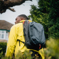 A person sporting a Cancha Padel & Pickleball Bundle's yellow jacket and large black backpack with modular accessories strolls near trees and a windowed building.