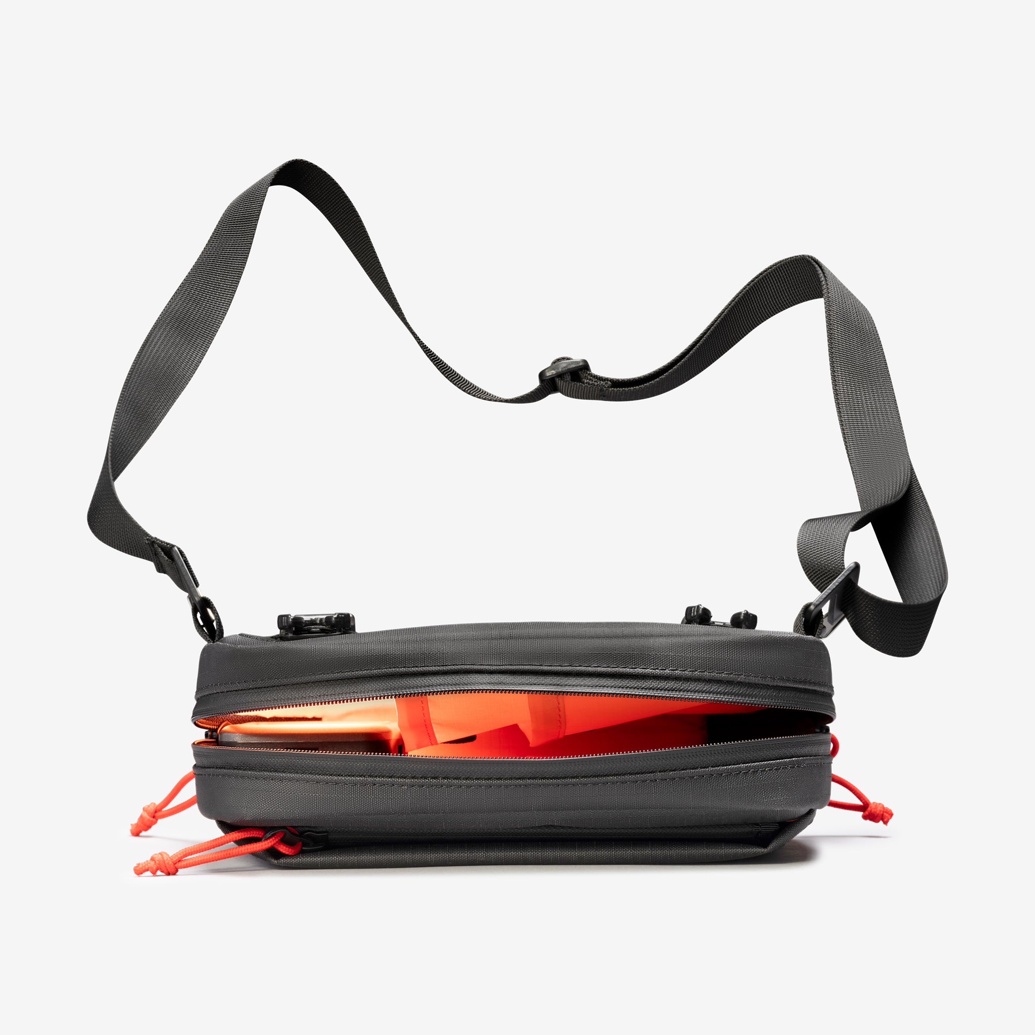 A Cancha Sling Bag, featuring a sleek black design and a convenient shoulder strap, is shown partially unzipped to reveal orange items inside. The versatile bag includes weather-resistant pockets and distinctive red zipper pulls.