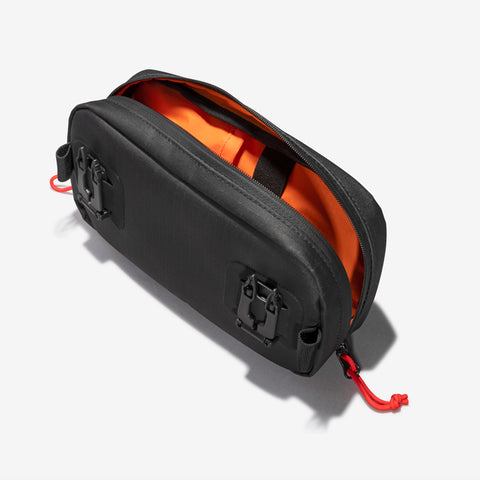 The Cancha Sling Bag is a black rectangular zippered pouch with an open top revealing a bright orange interior, featuring red zipper pulls, black clasps on the front, and weather-resistant pockets.