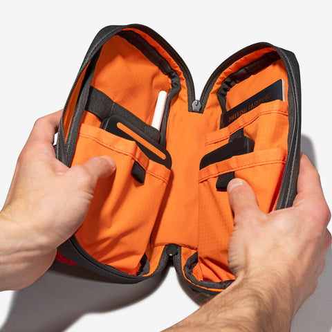 Two hands hold open a small, orange, zippered pouch with multiple internal pockets, revealing various organized tools inside. Its versatile design ensures travel functionality, making the Cancha Sling Bag an essential companion for any adventure.