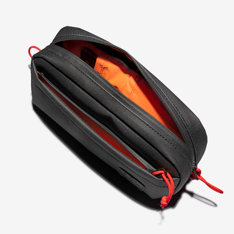 A Cancha Sling Bag, black with an orange interior and red pull tabs, is open to reveal two weather-resistant pockets inside.