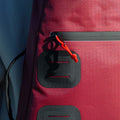 Close-up image of The Original Racquet Bag (15L) by Cancha in maroon, featuring a zippered pocket with an orange zipper pull. The grid-patterned fabric includes black attachment points below the zipper. This lightweight, water-repelling pack easily fits essentials and offers customizable add-ons for convenience.