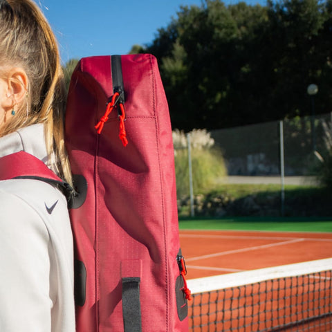 A person with blonde hair is facing away, wearing a white jacket and carrying "The Original Racquet Bag (15L)" by Cancha. The bag, made from lightweight water-repelling material and offering customizable add-ons, is slung casually over their shoulder as they stand near a tennis court.