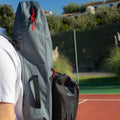 Close-up of a person carrying a lightweight, water-repelling gray backpack and The Original Racquet Bag (15L) by Cancha, which looks like it could be a tennis racquet bag, while standing next to a tennis court on a sunny day.