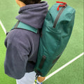 A person wearing a dark hoodie is carrying "The Original Racquet Bag (15L)" by Cancha, which is a large green backpack with red zippers, on a green outdoor surface reminiscent of a tennis court.